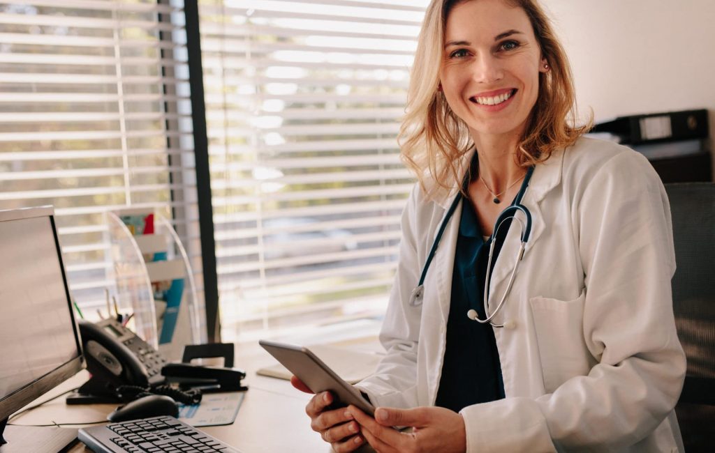 Friendly female doctor at her clinic desk Female doctor sitting at her desk holding a digital tablet. Positive female physician looking at camera with smiling on the face.