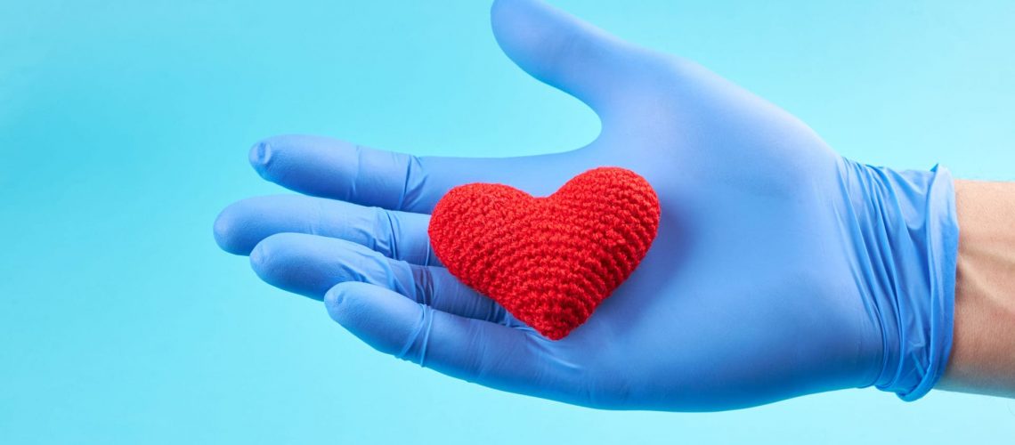 red heart symbol at persons hand  with blue medical gloves on. light blue background with copy space. heart disease concept.